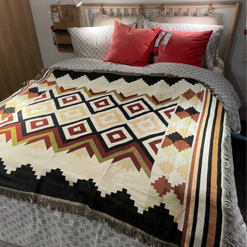 Dans une petite chambre, on voit un lit deux places avec une parure grise et des oreillers rouges surmontés d'un plaid aux motifs bohèmes, ethniques.