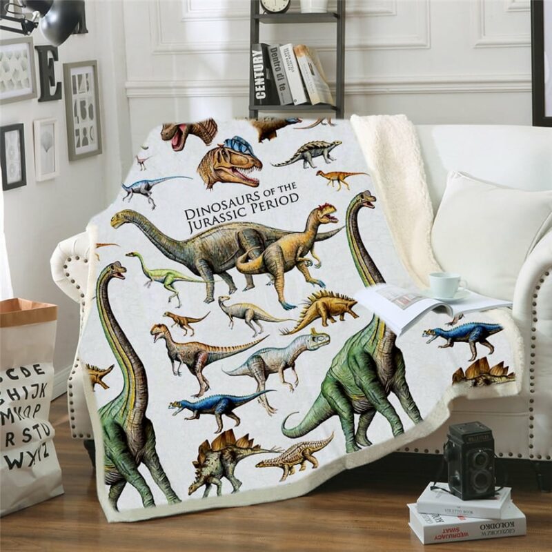 Plaid polaire blanc avec différentes espèces de dinosaures étendu sur un canapé blanc à côté d'un coussin blanc. Un livre ouvert avec une tasse est posé dessus. Deux livres sont sur le sol en parquet avec un appareil noir dessus .