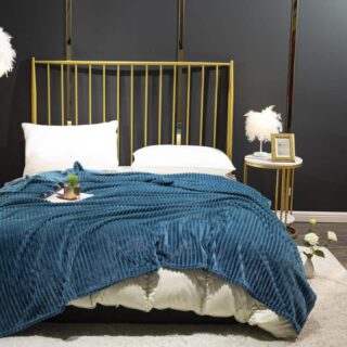 Couverture en flanelle côtelé bleu canard étendu sur un lit à la literie blanche et à la tête de lit à barreaux dorées. Le lit est posé contre un mur noir avec une table de chevet à côté.