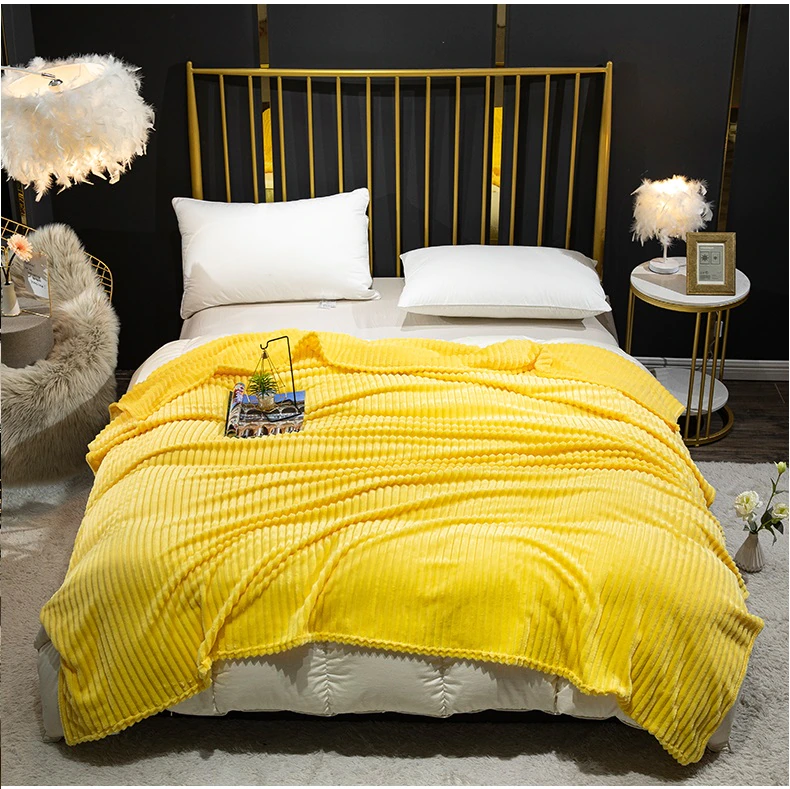 Plaid jaune polaire côtelé étendu sur une literie blanche sur un lit 2 places avec une tête de lit à barreaux dorés. Il y a une table de chevet et les murs de la chambre sont noirs.