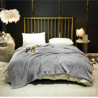 Couverture grise en velours côtelé étendue sur un lit deux places avec des coussins blancs, une tête de lit à barreaux dorés. La chambre à un mur noir avec un grand tapis au sol beige en fourrure et des lampes ornées de plumes.