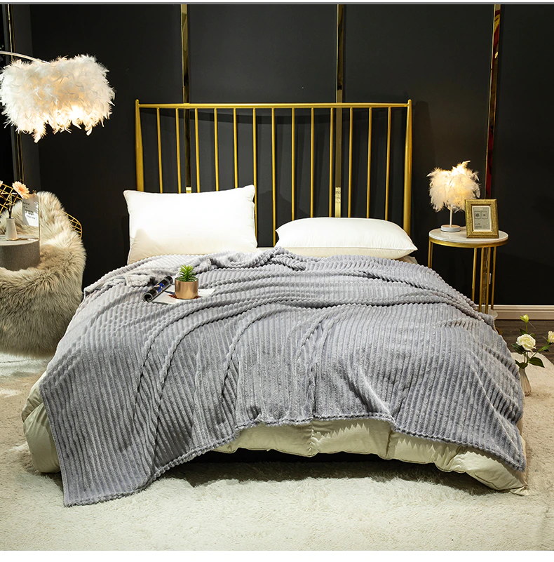 Couverture grise en velours côtelé étendue sur un lit deux places avec des coussins blancs, une tête de lit à barreaux dorés. La chambre à un mur noir avec un grand tapis au sol beige en fourrure et des lampes ornées de plumes.