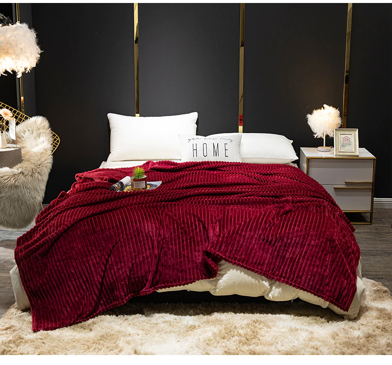Couverture en flanelle côtelé rouge étendue sur un lit à la literie blanche. Le lit est posé contre un mur noir avec une table de chevet à côté avec un tapis en fourrure beige en premier plan.