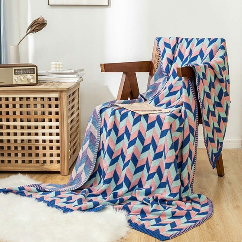 Couverture avec formes triangulaires bleu foncé, rose et bleu clair posé sur une chaise en bois débordant au sol sur un tapis blanc. Il y a un meuble en bois ajouré de petits carreaux avec une lampe et une vieille radio dessus.