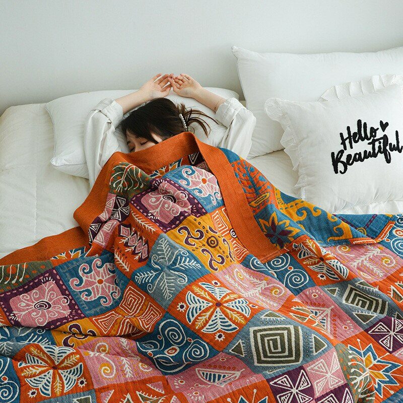 Une jeune fille brune allongée sur un lit blanc recouverte d'un plaid orangé à carreaux et motifs multicolores. Il y a un oreiller blanc avec inscription en noir "hello beautiful"