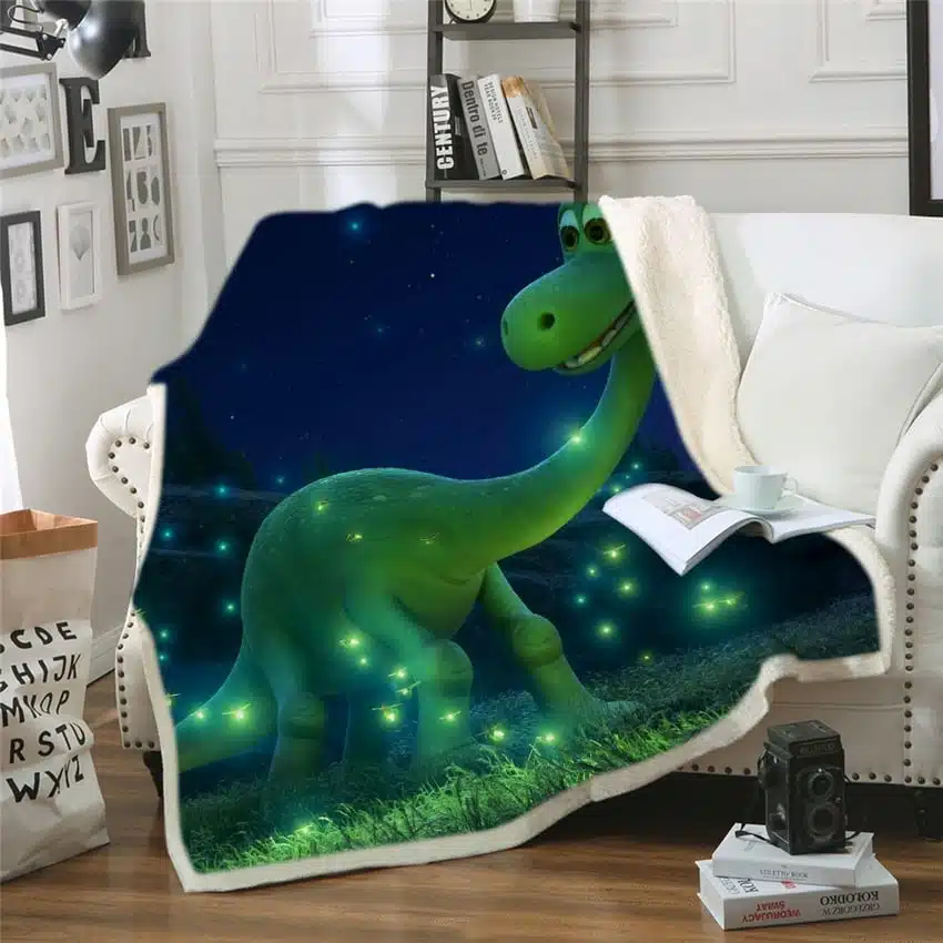 Couverture polaire bleue avec dinosaure de dessin animé vert, étendue sur un canapé à côté d'un coussin blanc. Il y a un livre ouvert avec une tasse posée dessus et deux livres au sol avec un appareil noir sur le dessus.