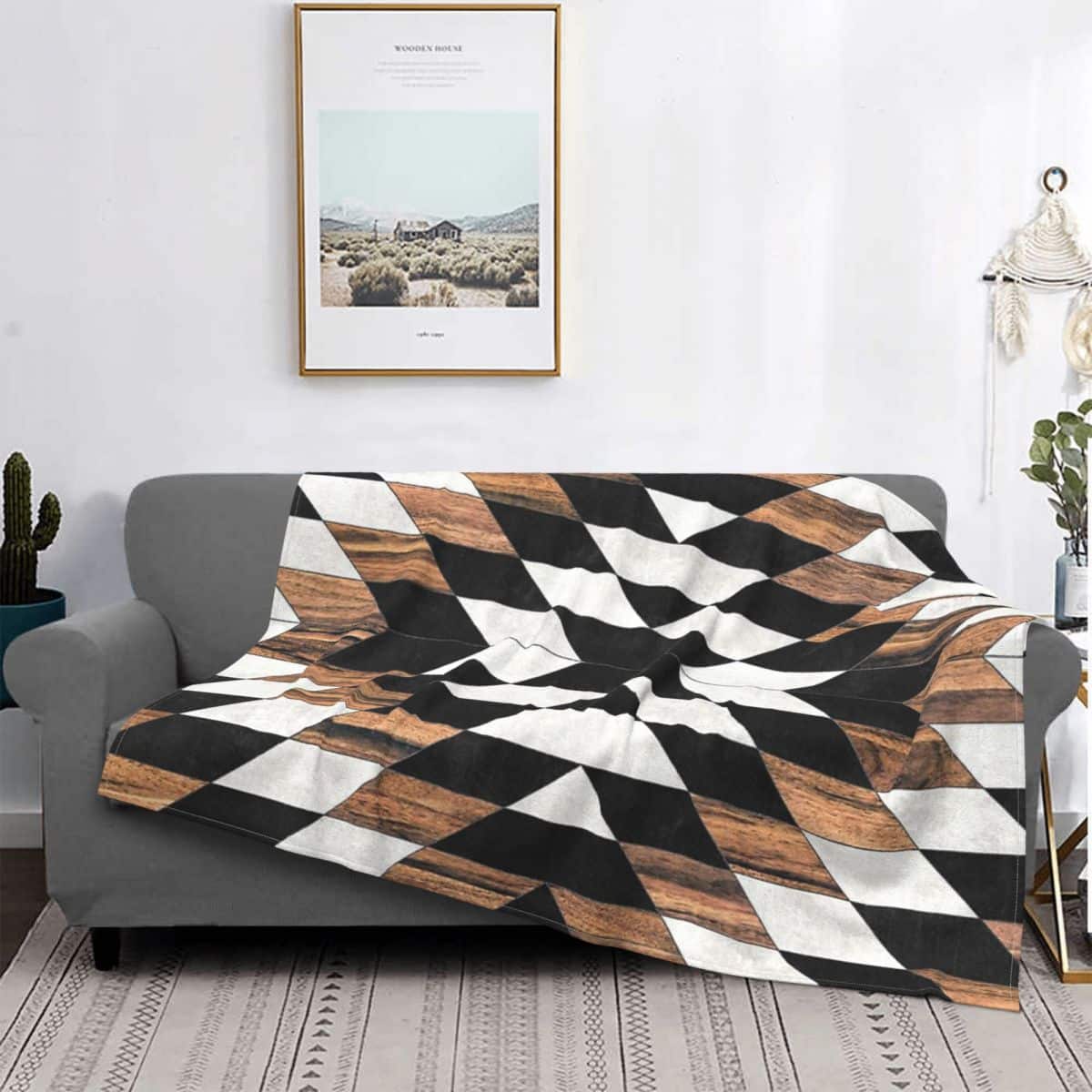 Plaid aux motifs tribal urbain blanc, marron et noir étendu sur un canapé gris. Il y a un cadre clair avec un paysage au dessus du canapé sur un mur blanc.
