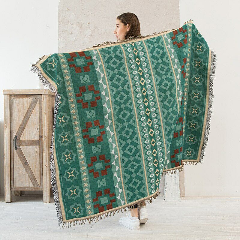 Femme de dos qui étend dans ses mains un plaid vert aux motifs géométriques qui lui couvre entièrement le dos dans une pièce blanche avec un meuble en bois .