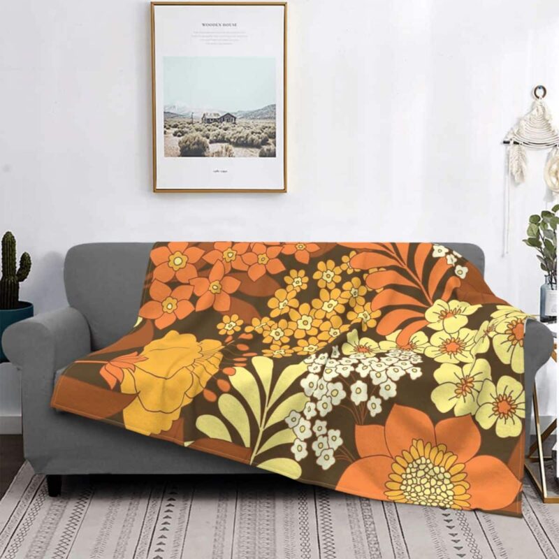 Plaid polaire vintage avec fleurs colorés en plusieurs nuances d'orange dans un style années 70 étendu sur un canapé gris dans un décor de salon. Le sol est un tapis beige au style bohème à rayures , il y a un cadre avec un paysage de nature au -dessus du canapé, un objet de déco en macramé accroché à coté et un petit cactus à l'opposé.