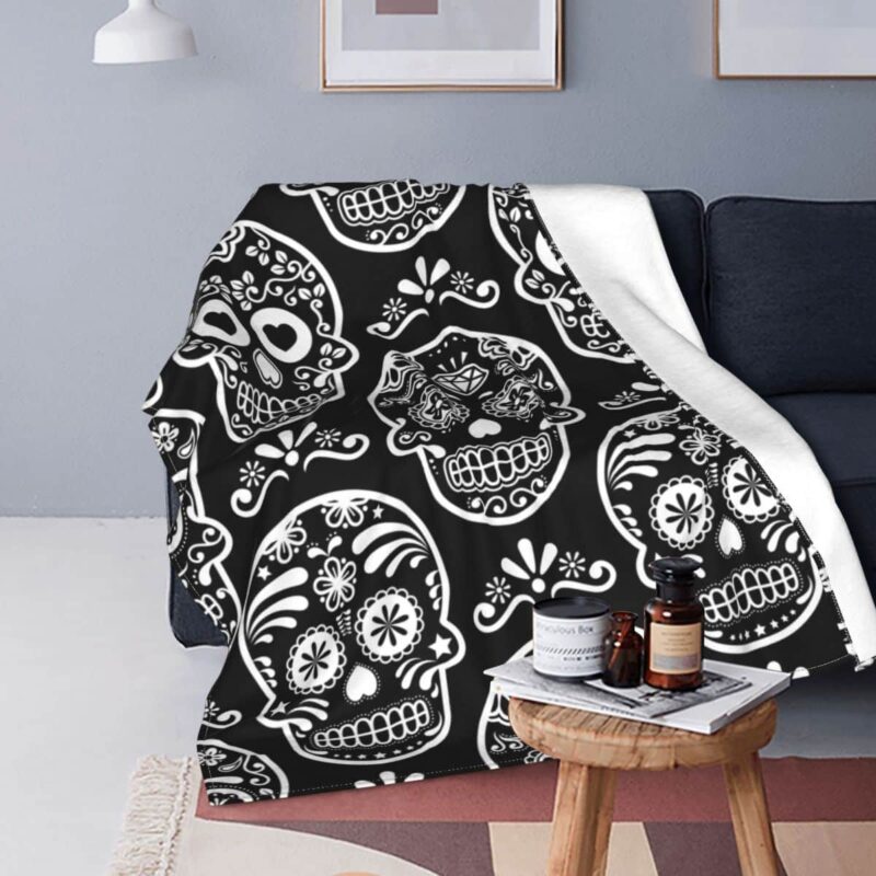 Plaid polaire noir et blanc avec têtes de mort mexicaines étendu sur un canapé noir avec une petite table d'appoint en bois ronde devant . Il y a un magazine posé dessus avec 3 pots en verre fumé.