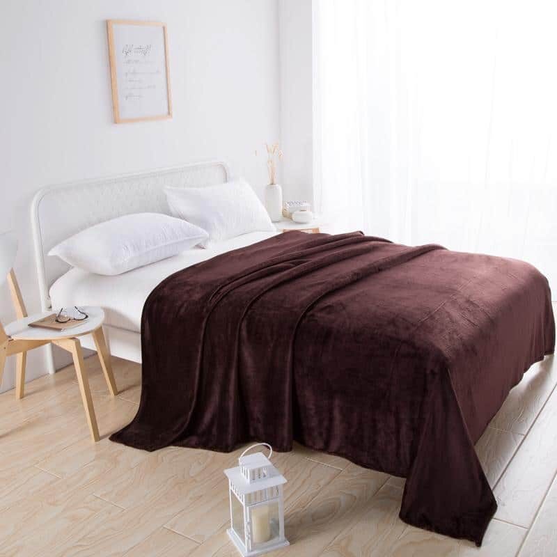 Plaid en velours marron couvrant un lit dans une chambre blanche au sol beige avec chaise en bois et lanterne blanche sur le sol