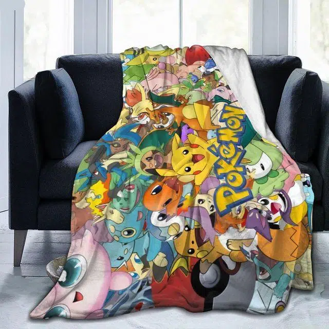 Plaid pokémon univers multicolore avec un medley de tous les principaux personnages. Il est étendu sur un fauteuil gris foncé et déborde sur le sol en parquet gris clair.