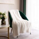 Plaid polaire fourrure blanc étendu sur un fauteuil en velours vert sur un tapis blanc avec une petite table d'appoint ronde ne marbre avec des fleurs blanches dans un vase posé dessus.
