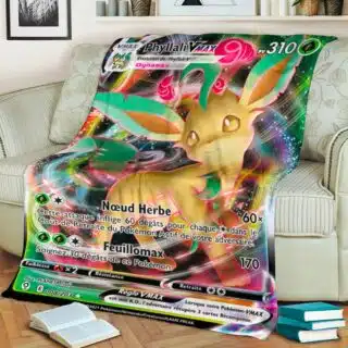 Plaid polaire multicolore carte Pokémon Phyllali Vmax, étendu sur un canapé beige fleuri et rayé et débordant sur le sol en parquet marron. Il y a une pile de 3 livres et un livre ouvert sur le sol.