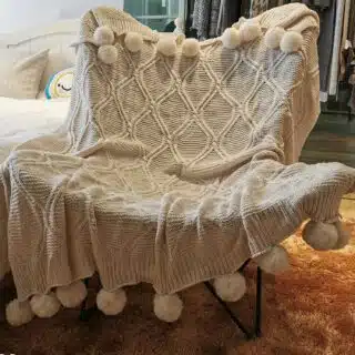 Plaid beige tricoté en acrylique avec pompons, posé sur une chaise devant une penderie et un tapis marron