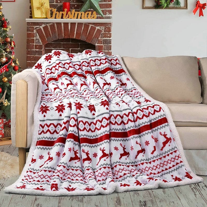 Plaid couverture rouge et blanc avec différents motifs de Noël comme le cerf dessus, posé sur un canapé beige et devant une cheminée et un sapin de noël avec des décorations or et rouge