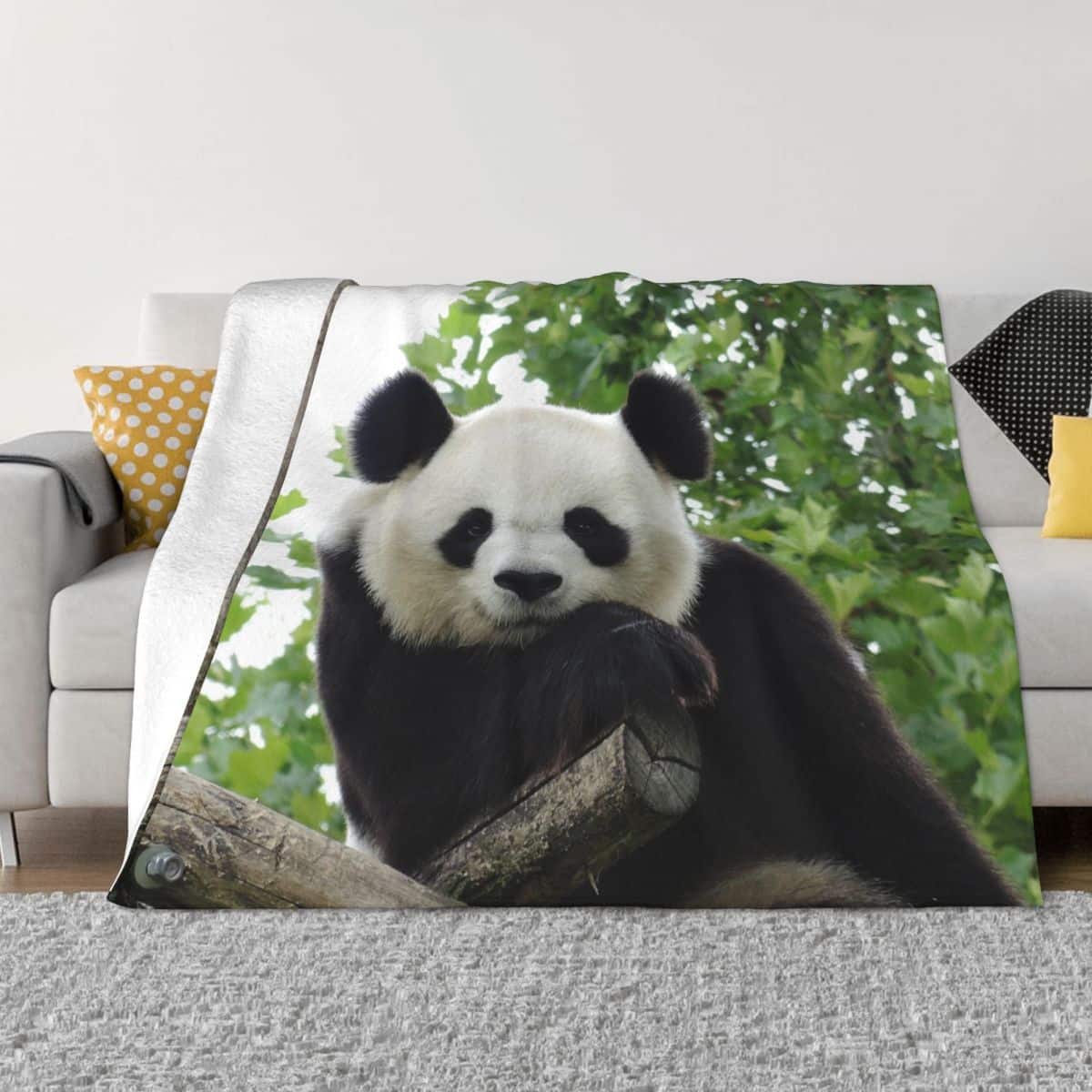 Plaid en flanelle avec un motif de Panda blanc et noir devant des feuilles vertes, posé sur un canapé gris avec des coussins de couleurs jaunes