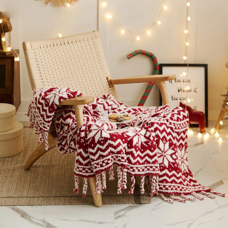 Plaid tricoté de noël rouge et blanc posé sur une chaise beige avec une assiette avec des biscuits posés dessus devant un mur blanc avec une guirlande lumineuse