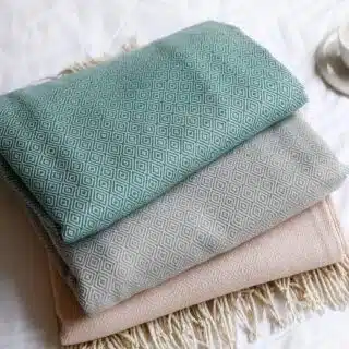 Plaid canapé à motif de losange de couleur vert, gris et rose à frange beige, plié et posé sur un drap blanc