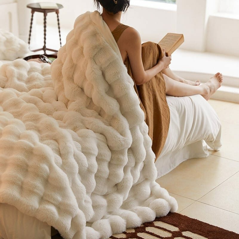 Plaid polaire blanc posé sur un lit et le dos d'une femme portant une robe marron et lisant un livre