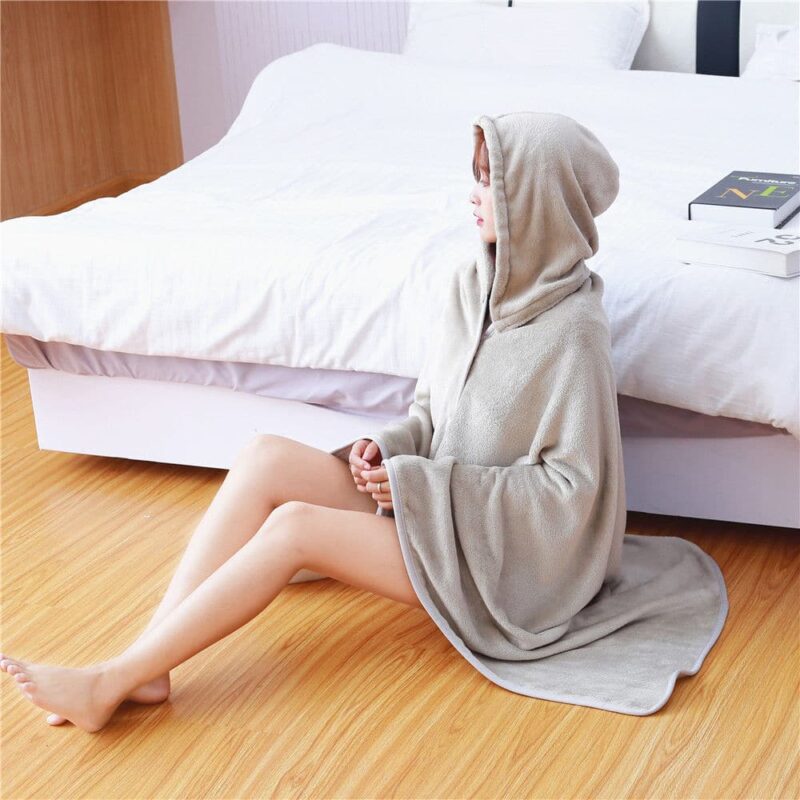 Pull plaid gris à capuche porté par une femme assise sur du parquet et adossée à un lit blanc