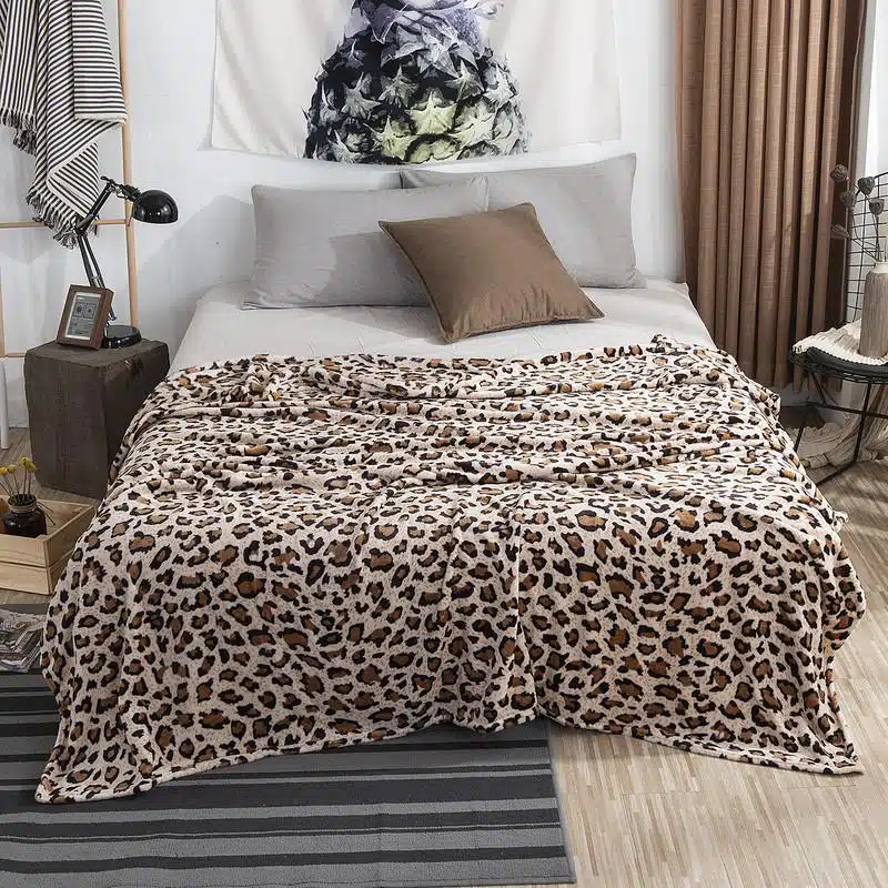 Plaid léopard de couleur beige, marron et noir, posé sur un lit aux draps blancs sur un sol en parquet et un tapis de couleur gris