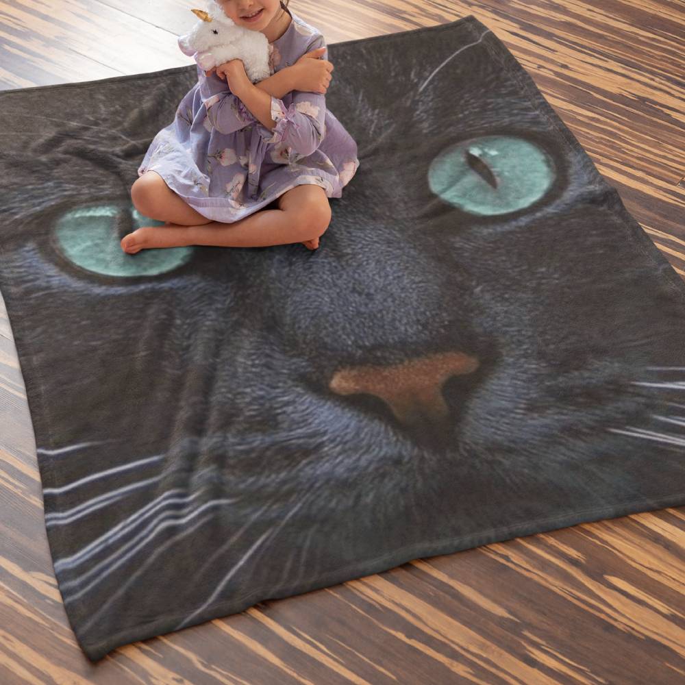 Une petite fille assise par terre sur un plaid avec une tête de chat noir vue de près et dont les yeux sont bleus et perçants