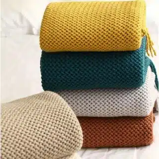 couverture tricot grosse maille couleurs tendances pliée et déclinée en jaune, vert, blanc, marron et beige, sur une table.
