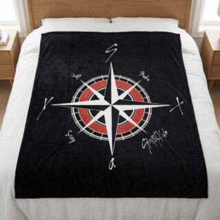 Un plaid Kpop boussole Stray Kids noir étendu sur un lit blanc, avec un grand dessin de boussole rouge et blanche au centre entourée d'inscriptions, dans une chambre lumineuse avec des tables de nuit en bois.